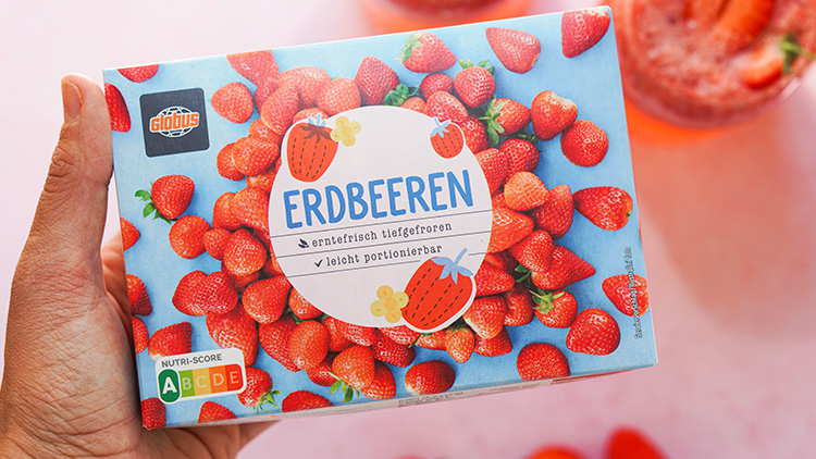 Eine Hand hält eine GLOBUS-Verpackung mit tiefgefrorenen Erdbeeren, NUTRI-SCORE A und blauem Hintergrund mit Erdbeermuster.