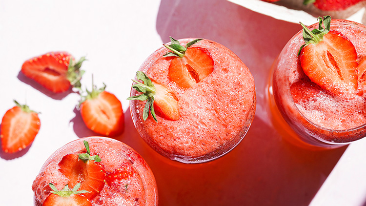 Drei Gläser mit schaumigem Erdbeersaft dekoriert mit halbierten Erdbeeren, daneben weitere Erdbeeren auf einer weißen Oberfläche.