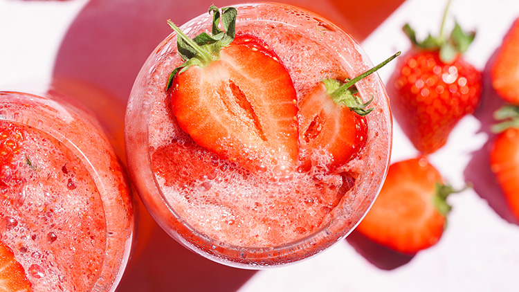 Ein Glas mit rotem Erdbeergetränk und frischen, halbierten Erdbeeren neben weiteren Erdbeeren auf weißem Untergrund.