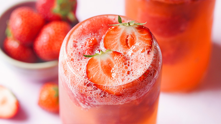 Ein Glas mit erfrischendem roten Getränk und Erdbeerscheiben, daneben unscharf Erdbeeren in einer Schale.