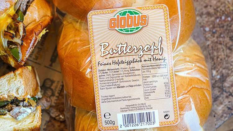 Eine eingeschweißte Packung GLOBUS Butterzopf, 500 g, mit Etikett und Inhaltsangaben liegt auf einem Tisch, daneben Sandwiches.