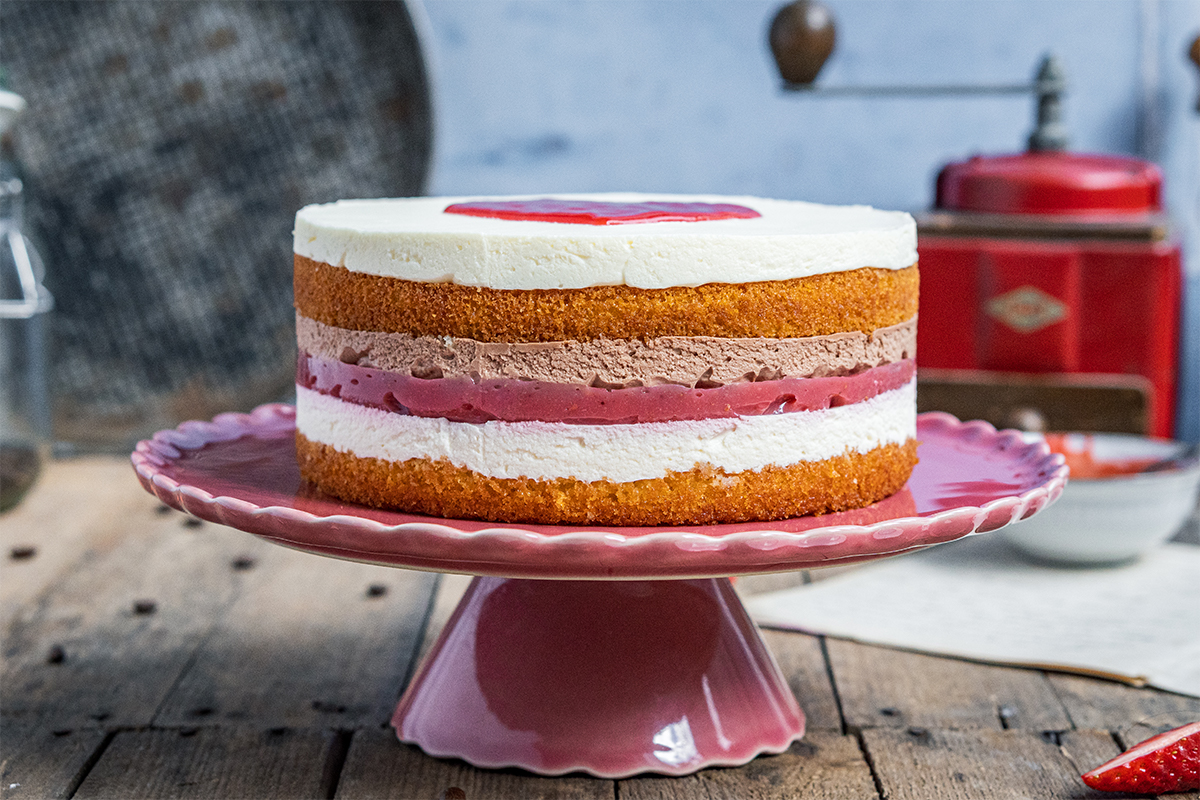 Schichttorte mit hellem und rotem Boden sowie Cremefüllung auf pinker Tortenplatte, im Hintergrund Küchenelemente.