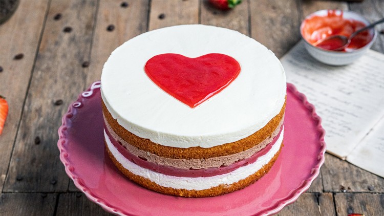 Erdbeer-Sahne-Torte mit Herz aus rotem Fruchtspiegel auf rosa Tortenplatte, Holzuntergrund, daneben Erdbeeren und Rezeptpapier.