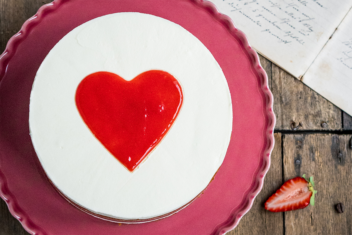 Eine Torte mit weißer Creme und rotem Herz aus Fruchtsoße auf rosa Teller, daneben eine halbierte Erdbeere und Papierseiten.
