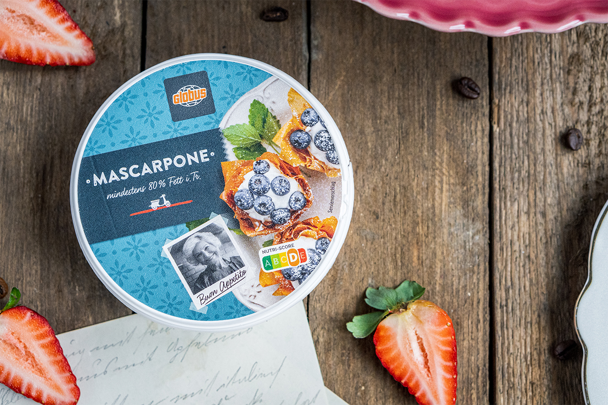 Runde Verpackung GLOBUS Mascarpone auf Holztisch, umgeben von Erdbeerhälften, Kaffeebohnen und einem beschrifteten Blatt Papier.