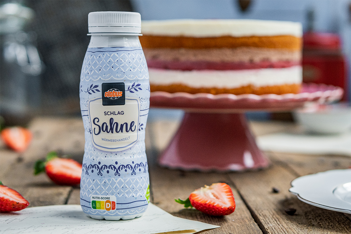 Eine Flasche GLOBUS Schlag-Sahne steht vor einem mehrschichtigen Kuchen auf einer rosa Tortenplatte, umgeben von Erdbeerstücken.