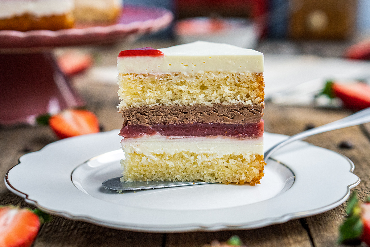 Ein Stück Erdbeer-Sahne-Torte mit Schichten aus gelbem Teig, Schokoladencreme und Erdbeergelee auf einem weißen Teller.