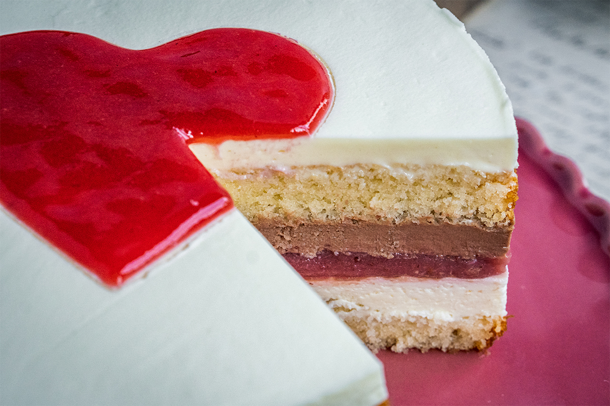 Torte mit rotem Fruchtspiegel in Herzform, innen Schichten aus heller Creme, Schokocreme, Fruchtgelee und Biskuit.