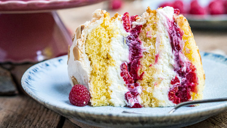 Ein Stück Biskuitkuchen mit Sahne, frischen Himbeeren und knuspriger Baiserkruste auf einem blau-grauen Teller.