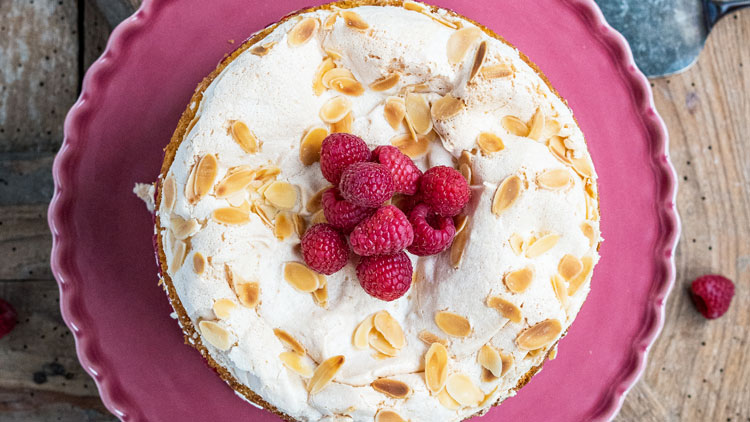Runder Kuchen mit heller Creme, gerösteten Mandelblättchen und frischen Himbeeren auf einer rosa Tortenplatte.