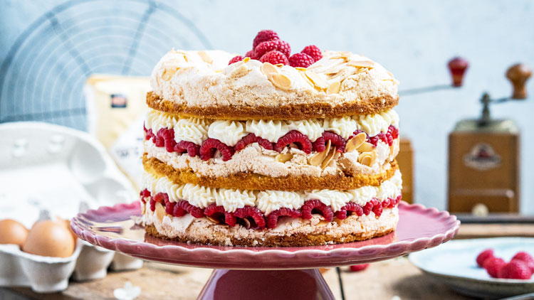 Eine mehrschichtige Himbeer-Baiser-Torte mit Sahne, Mandelblättchen und frischen Himbeeren auf einer rosa Tortenplatte.