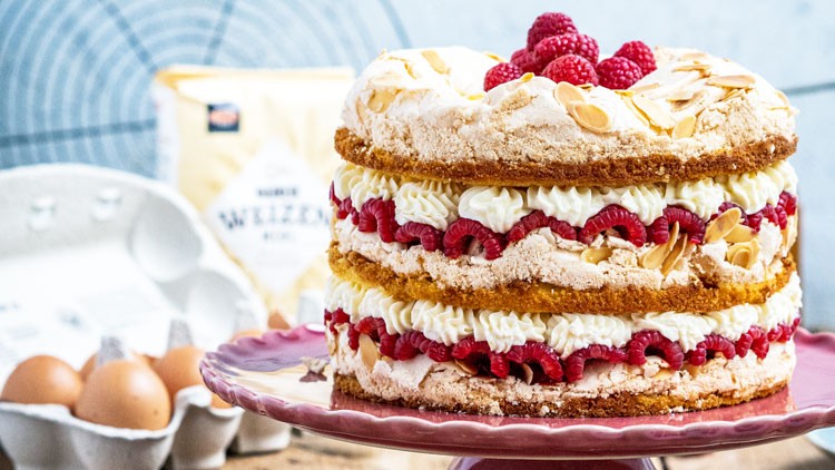 Mehrstöckige Himbeertorte mit Sahnecreme, Mandelsplittern und Himbeeren auf einem rosa Tortenständer, im Hintergrund Eier und Mehl.