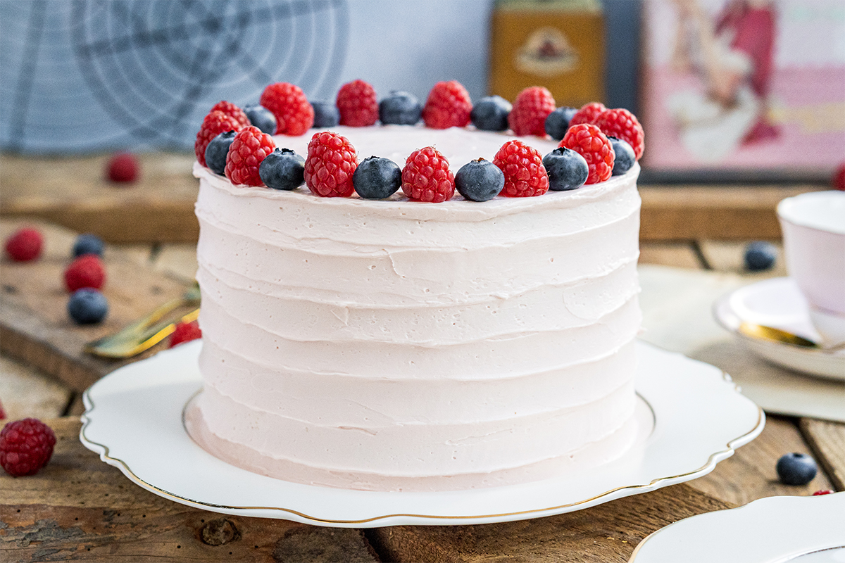 Runde Torte mit heller Creme und Dekoration aus Himbeeren und Blaubeeren auf einer weißen Servierplatte.