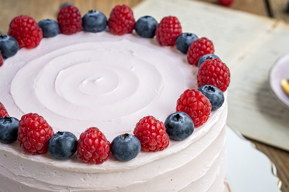 Runde Torte mit rosa Creme und Deko aus Himbeeren und Blaubeeren am oberen Rand auf einem Holztisch.