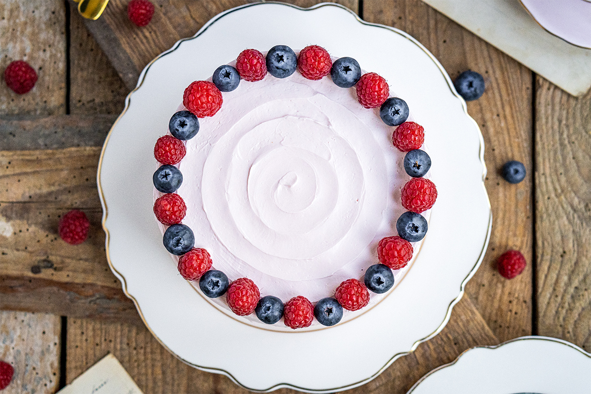 Runde Torte mit rosa Creme und einem Rand aus abwechselnden Himbeeren und Blaubeeren auf weißer Platte mit Holzuntergrund.