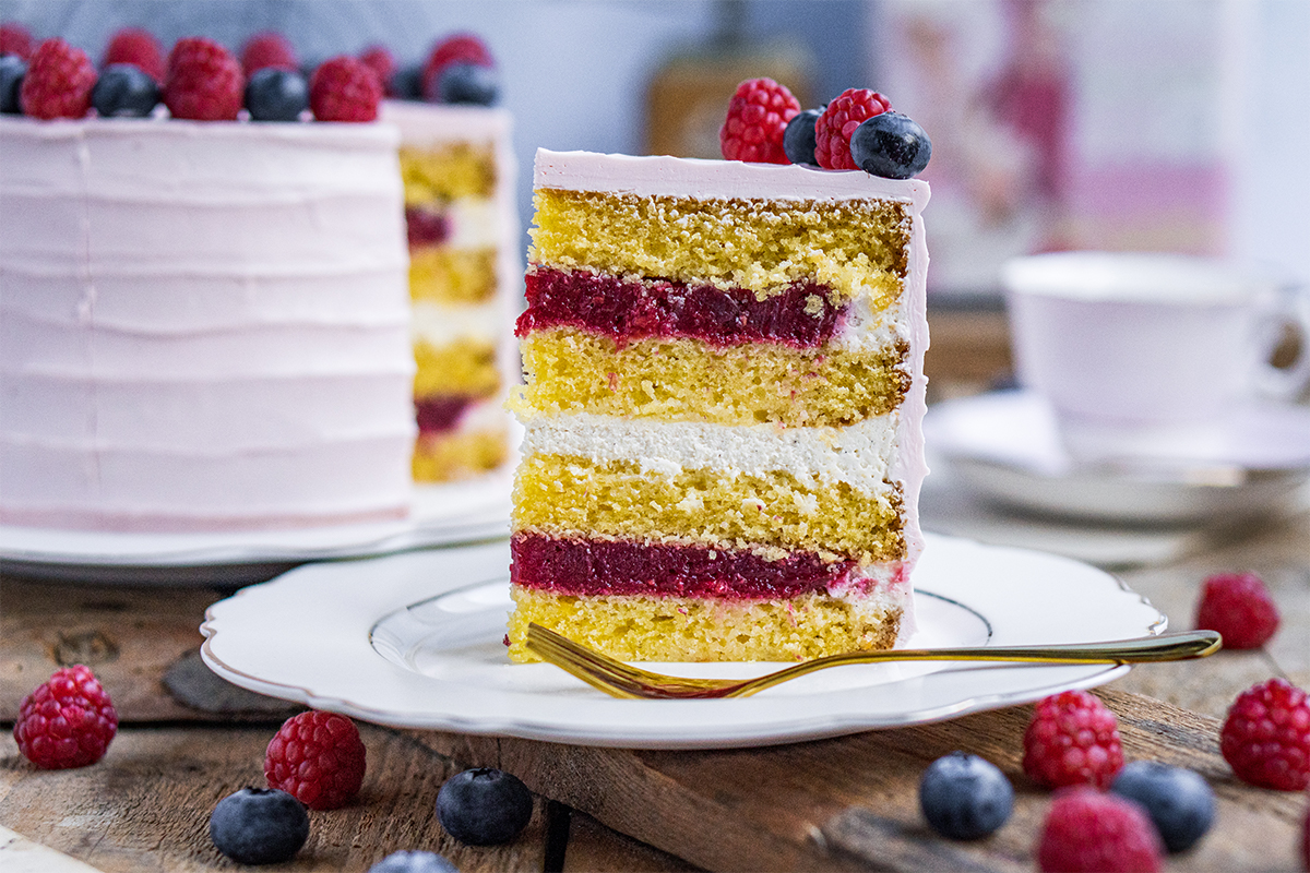 Ein Stück Torte mit hellrosa Creme, gelben Böden, roten Fruchtschichten und weißen Cremeschichten liegt auf einem Teller.