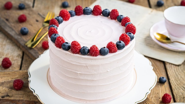 Runde Torte mit rosa Creme, dekoriert mit Himbeeren und Heidelbeeren, auf weißer Tortenplatte mit goldfarbenem Rand.