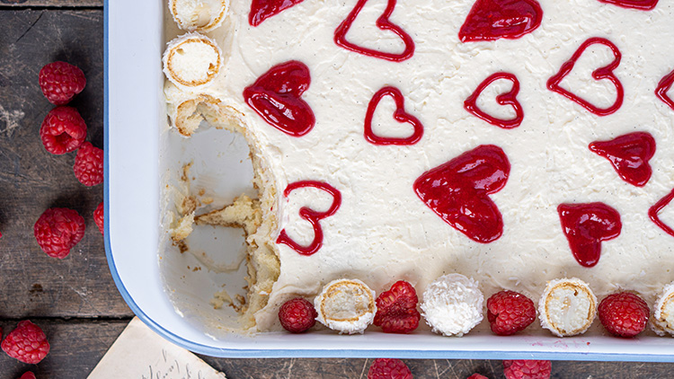 Eine Auflaufform mit Dessert, verziert mit roten Herzmotiven und Himbeeren, am Rand liegen Baiser und frische Himbeeren.
