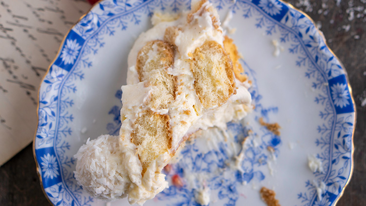 Ein Stück Tiramisu mit heller Creme und Löffelbiskuits liegt auf einem weiß-blauen Teller mit floralen Mustern.