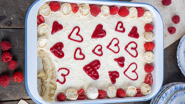 Rechteckige Auflaufform mit weißer Creme, roten Himbeeren und Herzmustern aus roter Soße, eine Ecke ist bereits entnommen.