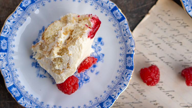 Ein Stück Sahnetorte mit Himbeeren liegt auf einem Teller mit blau-weißem Blumenmuster, daneben alte handgeschriebene Seiten.