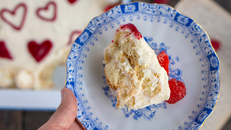 Ein Stück Kuchen mit heller Creme und Himbeeren liegt auf einem blau-weiß gemusterten Teller, darunter eine Hand.