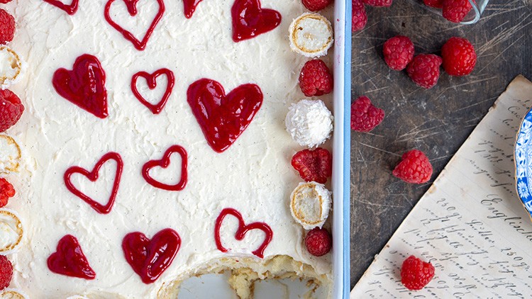 Eine helle Cremespeise in einer Form mit roten Himbeerherzen, frischen Himbeeren und weißen Konfektkugeln dekoriert.