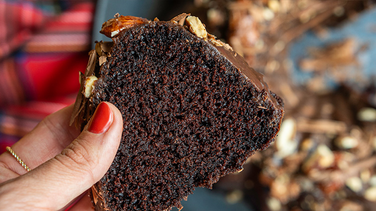 Ein Stück dunkler Schokoladenkuchen mit Nussstückchen wird in einer Hand mit rot lackierten Fingernägeln gehalten.