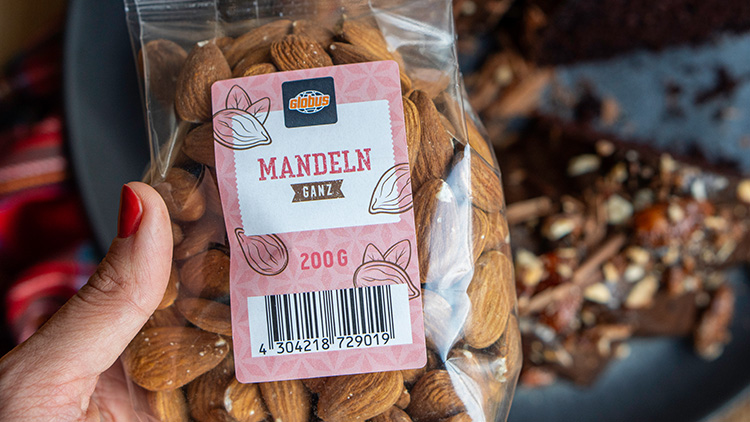 Eine Hand hält eine transparente Packung GLOBUS Mandeln ganz 200 g vor einem Teller mit Schokoladenkuchen.