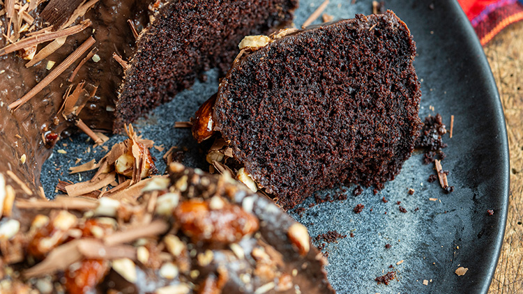 Ein angeschnittener dunkler Schokoladenkuchen mit Schokospänen und Nüssen auf einem dunklen Teller.