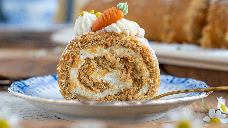 Ein Stück Karotten-Biskuitrolle mit Sahne, Marzipankarotte und gelben Streuseln auf einem Teller mit blauem Rand und goldener Gabel.
