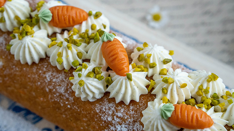 Rüblikuchen mit Puderzucker, Sahnetupfen, Marzipankarotten und gehackten Pistazien auf einer hellen Unterlage.