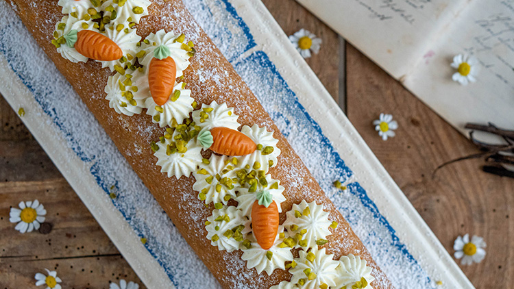 Eine Biskuitrolle mit Puderzucker, Sahnedekor, Marzipankarotten und gehackten Pistazien liegt auf einem blauen Teller.