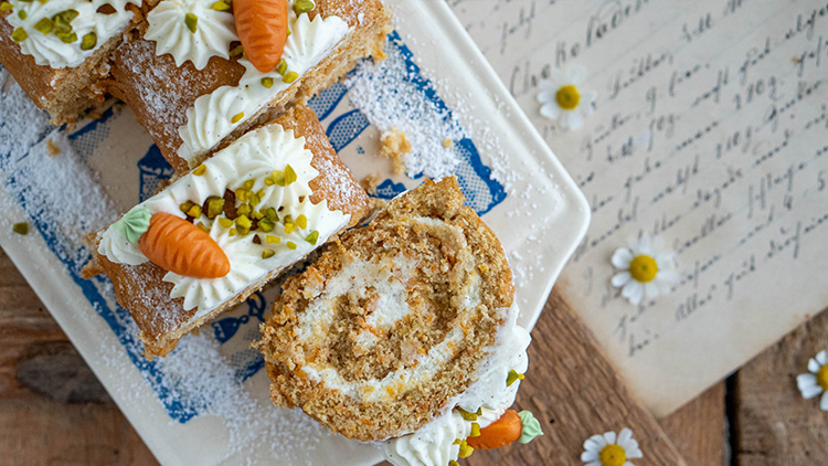 Karotten-Biskuitrolle mit Sahne, Marzipankarotten und Pistazien, dekoriert auf einem Teller neben handgeschriebenen Notizen.
