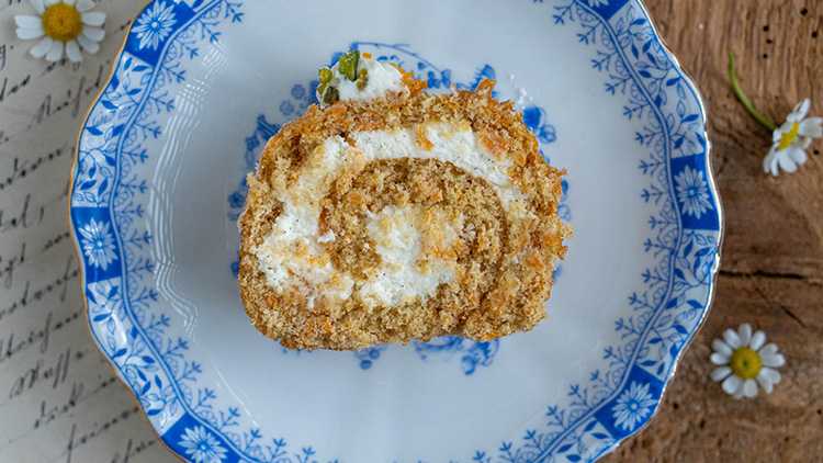 Scheiben einer dünnen Biskuitrolle mit heller Creme liegen auf einem weiß-blauen Teller, dekoriert mit kleinen Blüten.