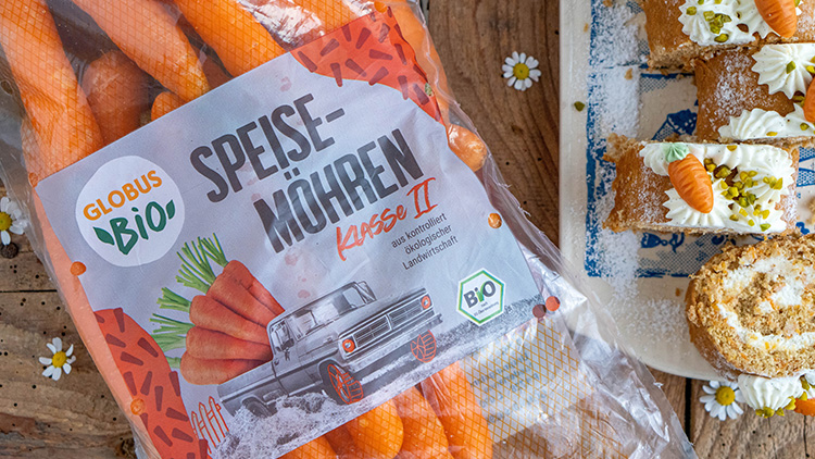 Eine Tüte GLOBUS Bio Speisemöhren Klasse II liegt neben Karottenkuchenstücken mit Sahne und Pistazien auf einem Holztisch.
