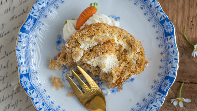 Ein angebissenes Stück Karottenkuchen mit Creme auf einem blauen Teller, daneben liegt eine goldfarbene Gabel.