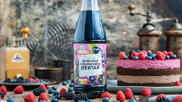 Eine Flasche GLOBUS Schwarzer Johannisbeer Nektar steht vor einer Beerentorte und frischen Beeren auf einem rustikalen Tisch.