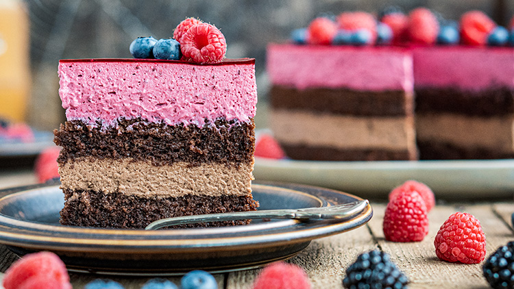 Ein Stück Schichttorte mit Beerencreme und Schokoladenschichten, dekoriert mit Himbeeren und Heidelbeeren auf einem Teller.