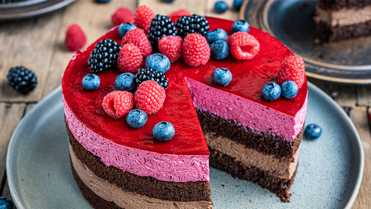 Runde Torte mit Schichten aus Schokolade und Beerencreme, belegt mit Himbeeren, Brombeeren und Blaubeeren, Stück herausgeschnitten.