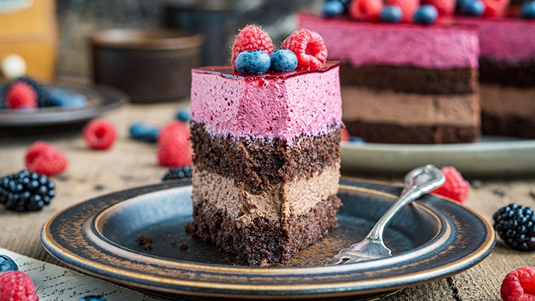 Ein Stück Schokoladen-Himbeer-Torte mit mehreren Cremeschichten und Beeren-Dekoration auf einem braunen Teller mit Gabel.