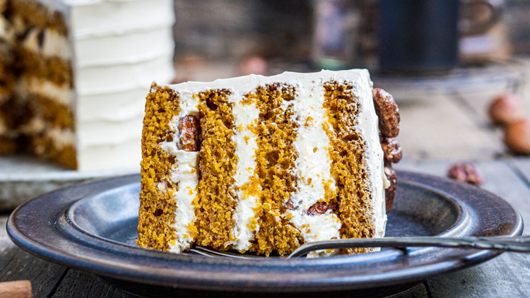Ein Stück Karottenkuchen mit weißer Cremeschicht liegt auf einem dunklen Teller, im Hintergrund eine Kaffeetasse.