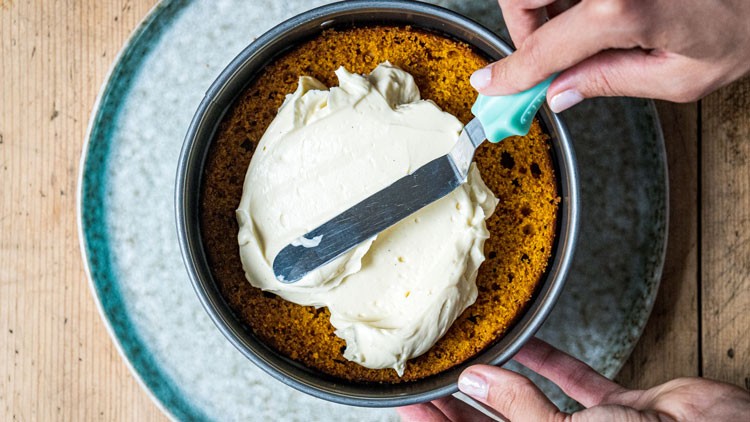 Eine runde Backform mit braunem Kuchenboden wird mit weißer Creme bestrichen, gehalten von zwei Händen.