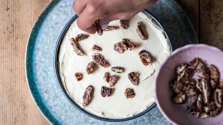 Eine Hand dekoriert eine helle Creme-Torte in einer runden Form mit karamellisierten Nüssen aus einer rosa Schale.