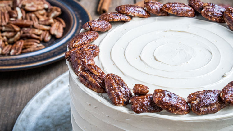 Runder Kuchen mit heller Creme und einem Rand aus glasierten Pekannüssen, daneben Teller mit weiteren Pekannüssen.