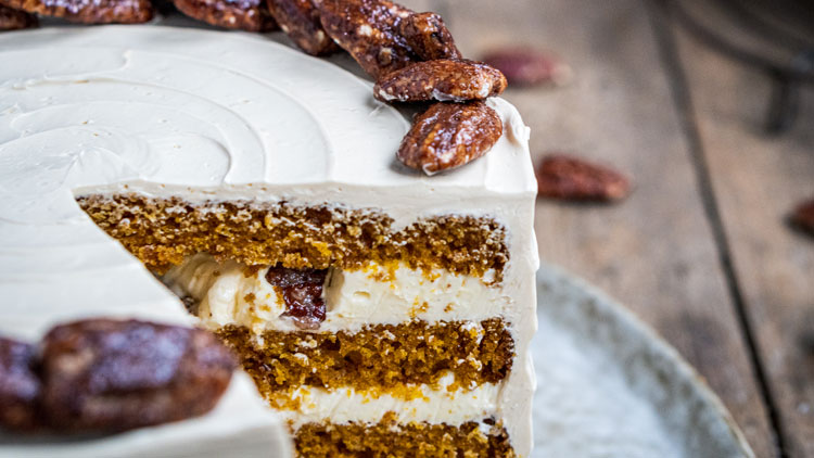 Anschnitt einer mehrlagigen Karottenkuchen-Torte mit heller Creme, dekoriert mit kandierten Nüssen auf einer Holzoberfläche.
