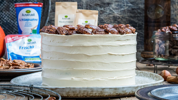 Weiße Torte mit Schichten aus Creme und kandierten Nüssen obenauf, umgeben von Backzutaten und Küchenutensilien.