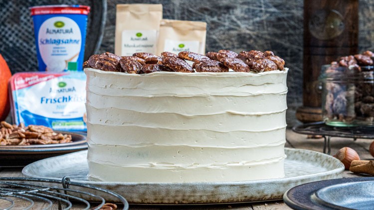 Weiße Torte mit Schichten aus Creme und kandierten Nüssen obenauf, umgeben von Backzutaten und Küchenutensilien.