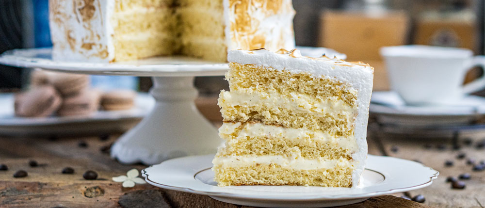 Ein Stück heller Schichtkuchen mit Sahnefüllung steht auf einem Teller neben einer Torte auf einer rustikalen Holzoberfläche.