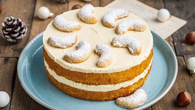 Runder Kuchen mit zwei Schichten Creme, dekoriert mit Puderzucker bestreuten Kipferln, auf blauem Teller und Holztisch.